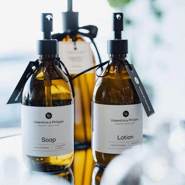 Drei elegante braune Glasflaschen der Naturkosmetiklinie „Valentina & Philippa“ mit schwarzem Pumpspender und weißem Etikett, dekorativ arrangiert auf einem Glastisch im Zimmer.